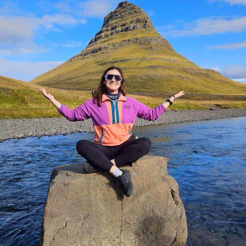 Kobieta siedzi na kamieniu nad rzeką z widokiem na zieloną górę Kirkjufell na Islandii